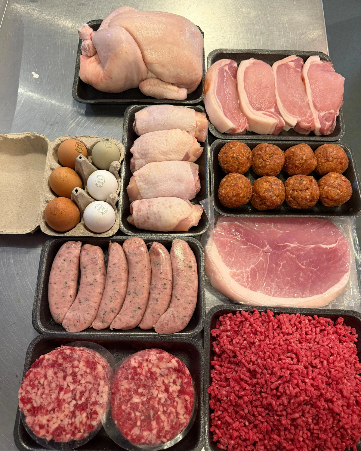 A variety of raw meat products arranged in black trays on a stainless steel surface, including a whole chicken, sliced pork chops, chicken thighs, meatballs, sausages, ground beef patties, and a large tray of ground meat.